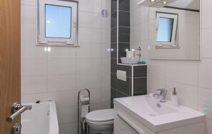 A modern bathroom with a white sink, toilet, and bathtub. There are two windows and a mirror above the sink. - Lovely apartment in Brela with WiFi (Brela)