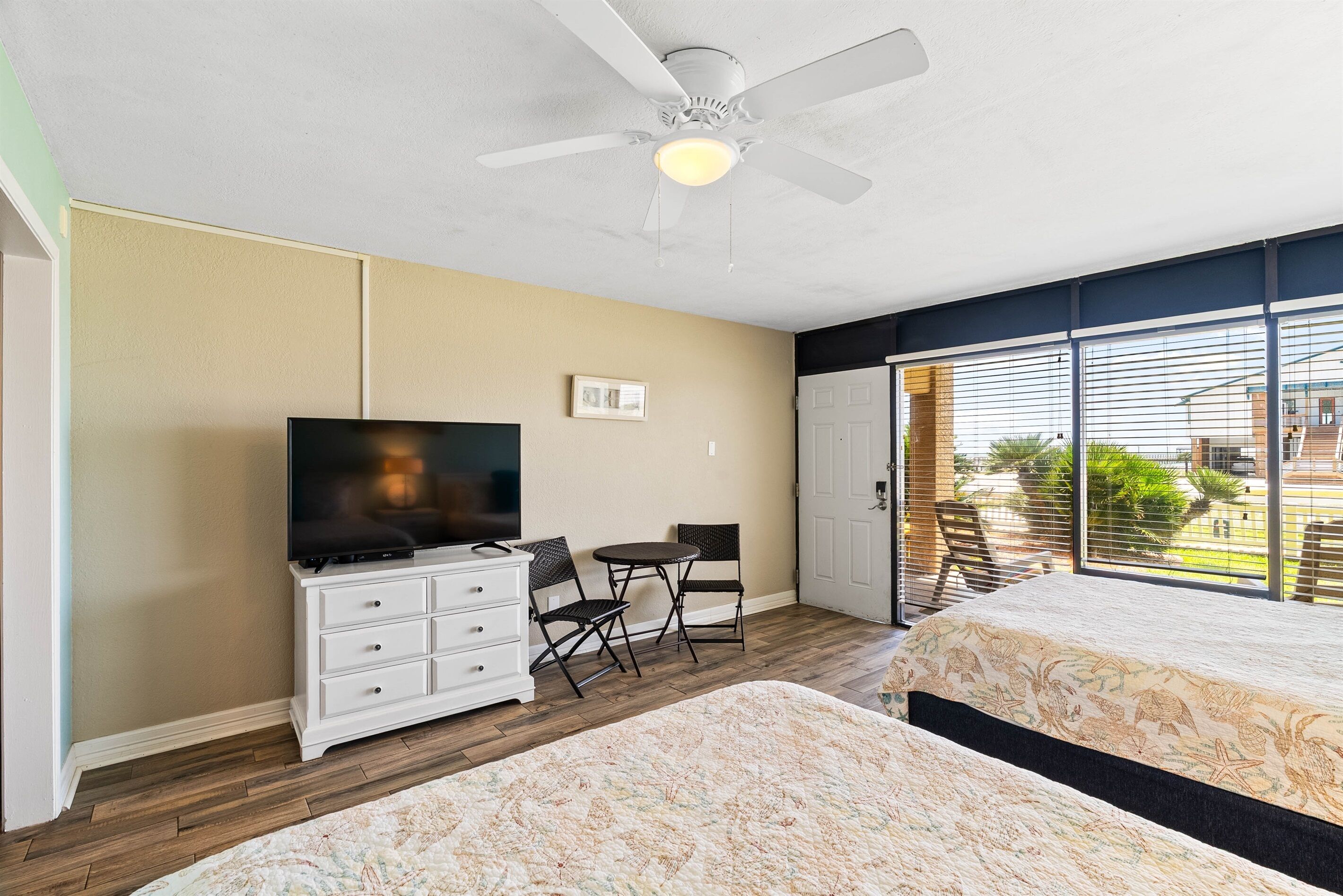 Habitación de hotel con cama, televisor, escritorio con sillas, ventilador de techo y ventana con persianas.