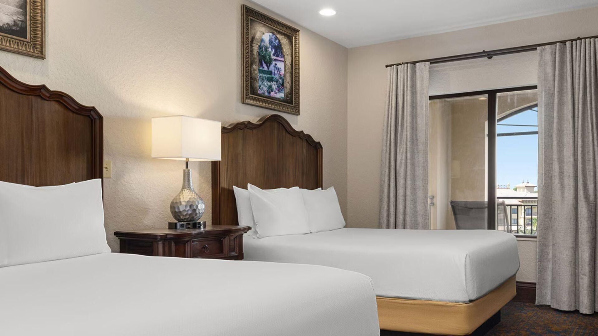 A hotel room with two beds, a nightstand with a lamp, a large window with curtains, and a framed picture on the wall.