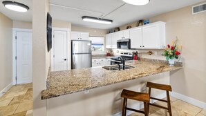 A modern kitchen with granite countertops, white cabinetry, stainless steel appliances, and a bar stool. - Welcome to Grandview – Laguna Reef Unit 414 (Rockport)