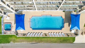 An aerial view of a hotel with a large rectangular swimming pool surrounded by sun loungers and umbrellas, a separate hot tub, and a fenced area with a small garden. - Welcome to Grandview – Laguna Reef Unit 414 (Rockport)