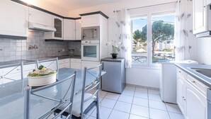 Eine moderne Küche mit weißen Schränken, einer eingebauten Mikrowelle, einem Backofen und einem kleinen Kühlschrank. Ein Esstisch mit Stühlen und ein Topf mit Pflanze stehen auf der Arbeitsfläche.