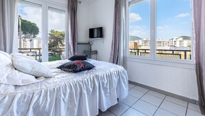 Ein Hotelzimmer mit Bett, Fernseher auf einer Ablage und großen Fenstern mit Vorhängen.