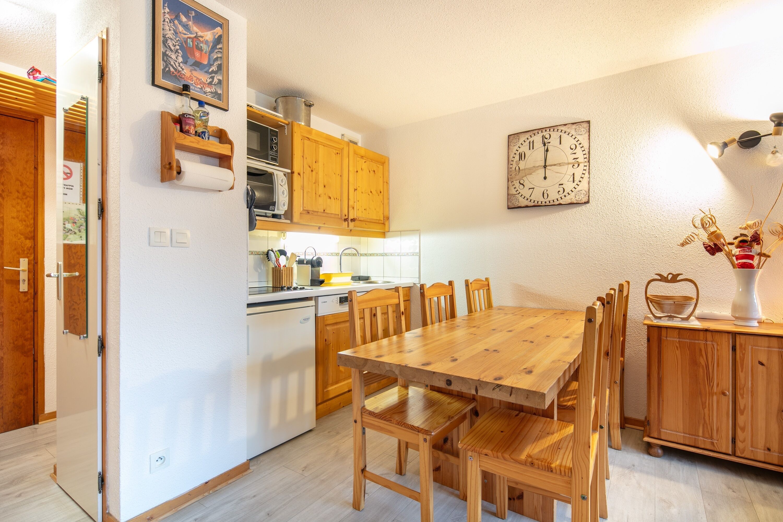 A kitchen with wooden cabinets, a wooden dining table with chairs, a clock on the wall, and a microwave.