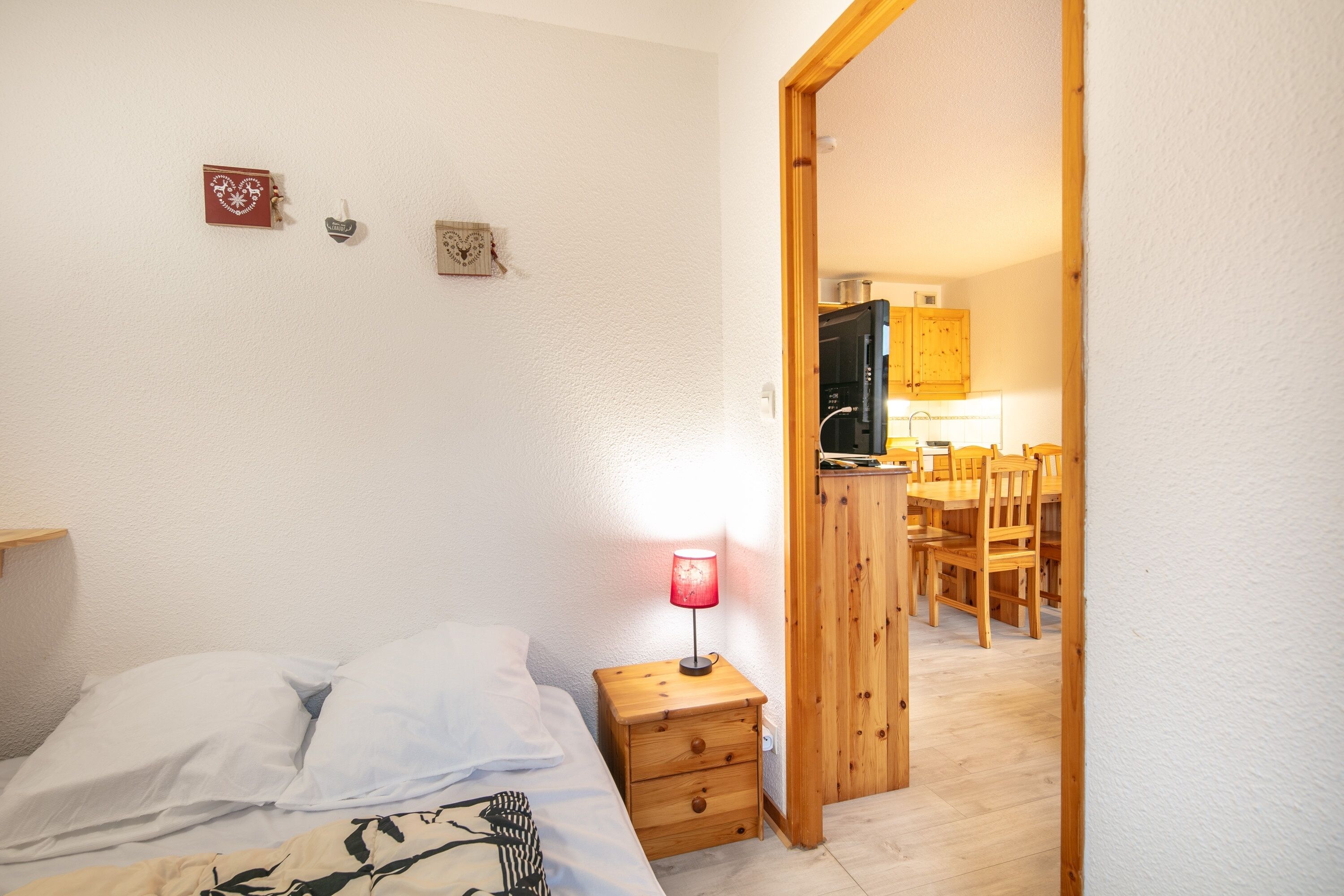 Une chambre à coucher comprenant un lit, une table de chevet et une lampe. Une porte en bois donne accès à un coin cuisine avec une table et des chaises.