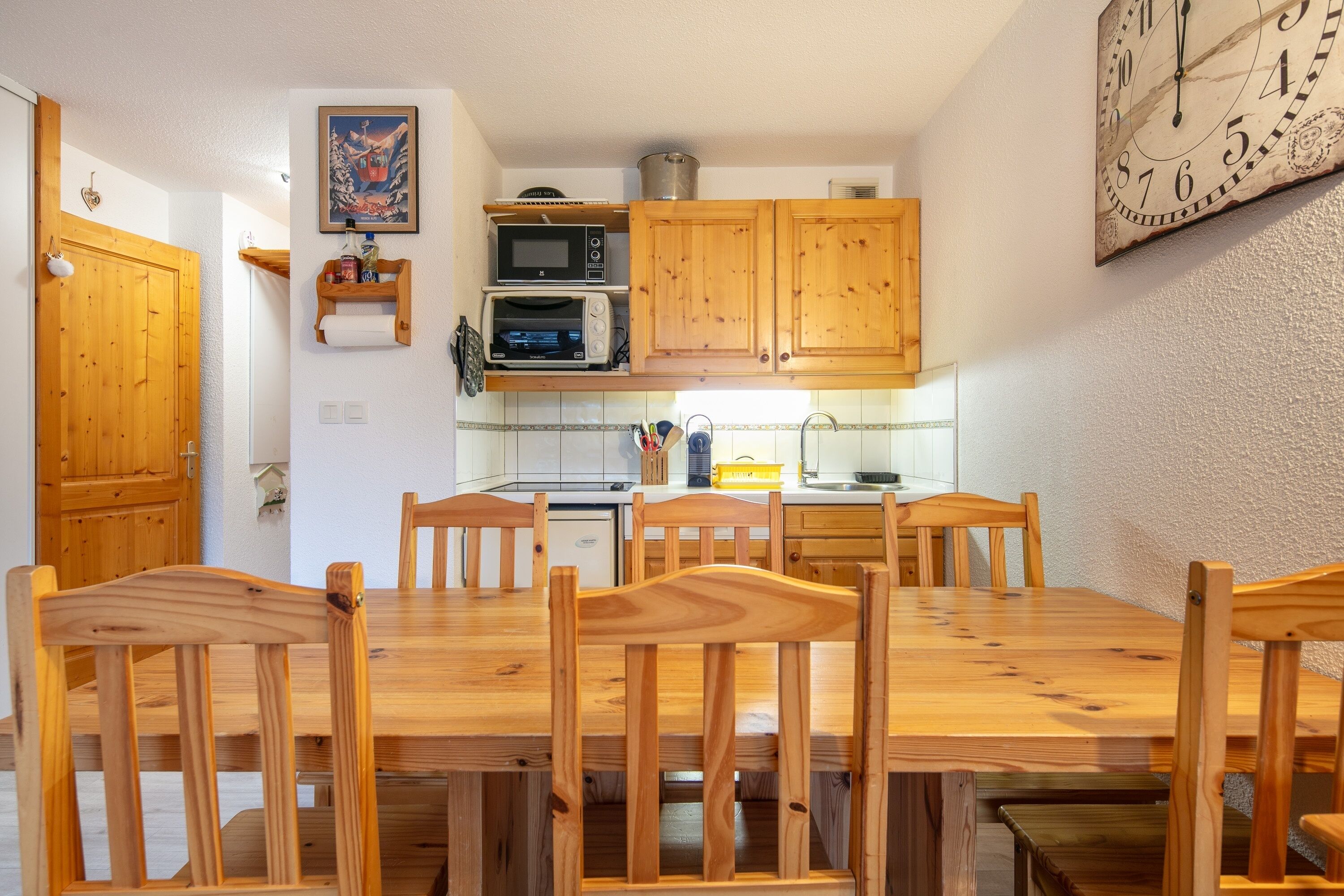A wooden dining table with six chairs, a kitchen with wooden cabinets, a microwave, and a clock on the wall.