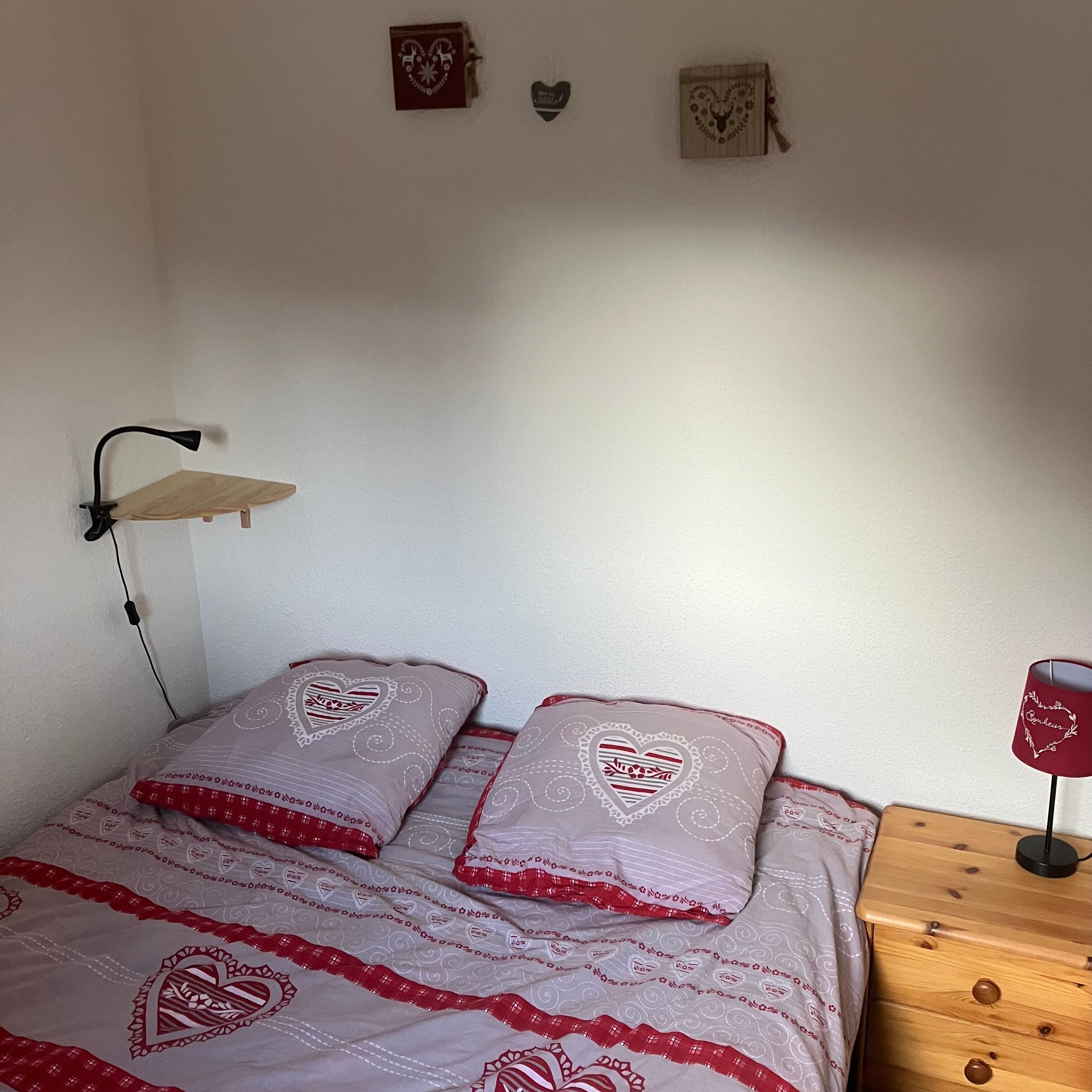 Ein Schlafzimmer mit einem Bett, zwei Kissen mit Herzmuster, einem Nachttisch mit Lampe und einem an der Wand befestigten Regal.