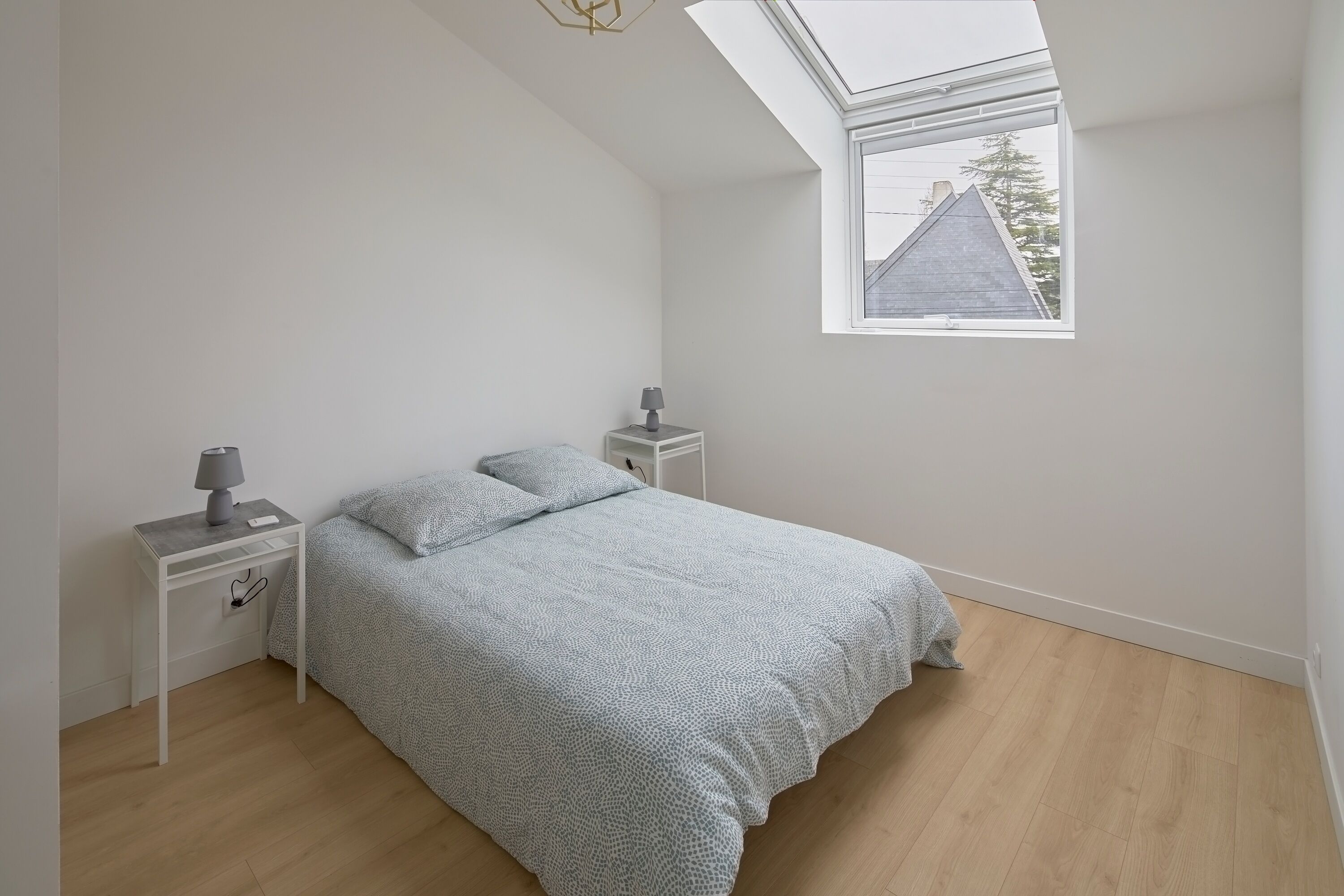 A bedroom with a skylight, a bed with a patterned bedspread, two bedside tables with lamps, and a view of a house through the window.