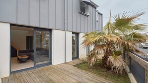 Uma casa moderna com um deck de madeira, porta de vidro que dá para o interior com um sofá e uma mesa, e uma palmeira no quintal.