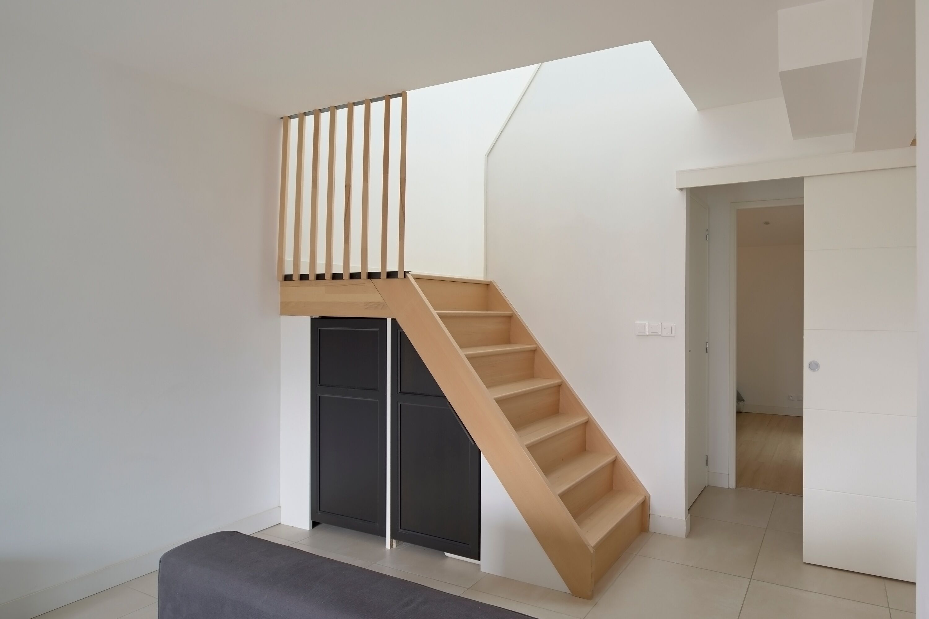 Un intérieur moderne avec un escalier en bois menant à une mezzanine, un banc et une porte donnant sur une autre pièce.