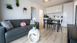 Un salon moderne avec un canapé gris, une table d’appoint blanche ornée d’une plante, et un coin repas comprenant une table et des chaises en bois.