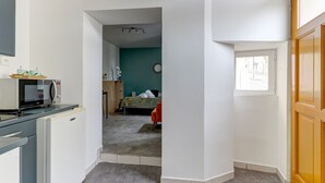 Een compacte keuken met een magnetron, een kleine koelkast en een spoelbak. Een doorgang leidt naar een slaapkamer met een bed en een stoel.