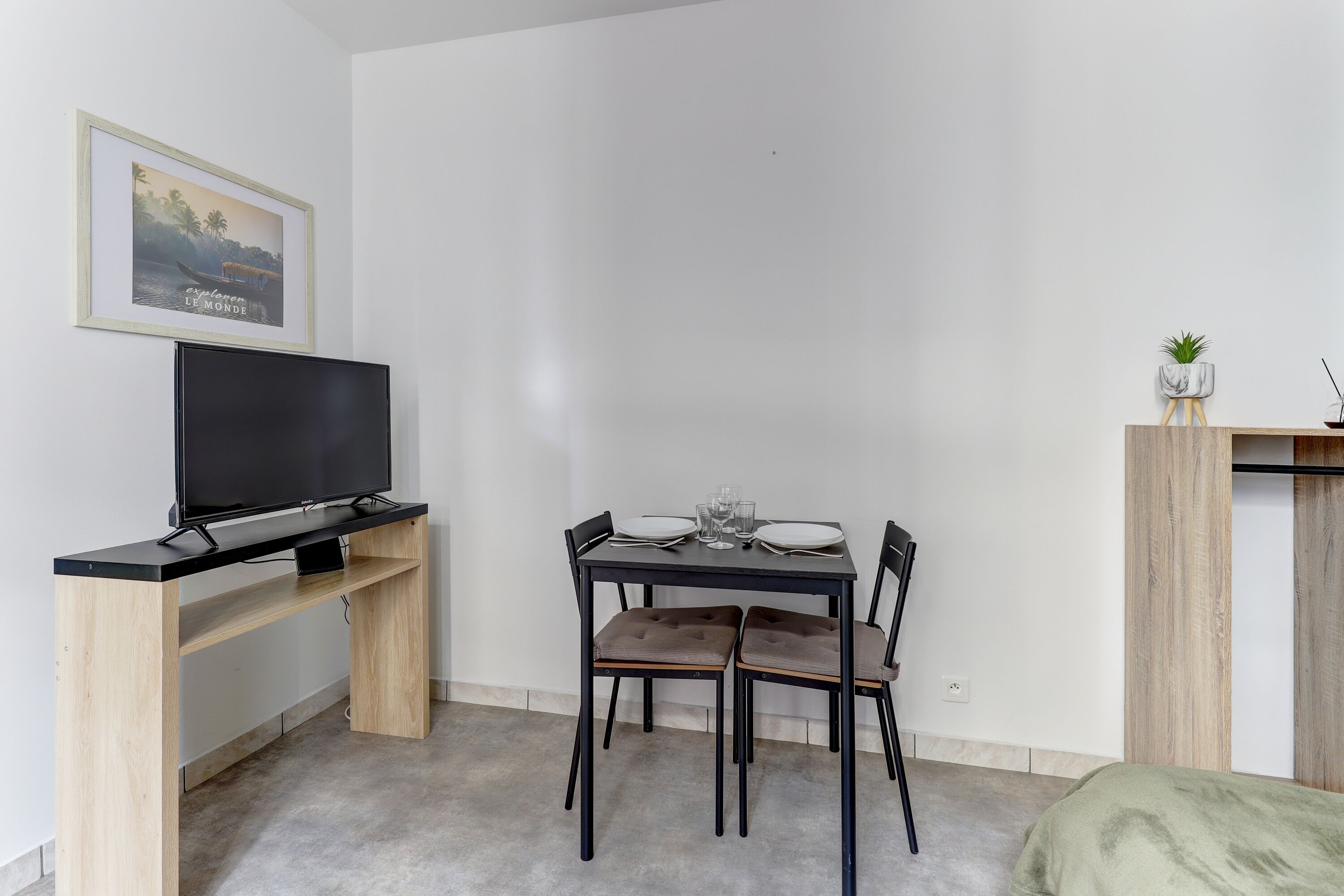 A compact living space with a wooden desk, a flat-screen TV, a dining table with chairs, and a small plant on a shelf.
