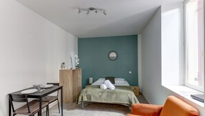 Een compacte kamer met een bed, een eettafel met stoelen, een klein bureau en een raam.