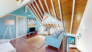 A spacious loft with a high ceiling, wooden beams, a large window, and a cozy seating area with a television and a painting. - Beautiful beach apartment with sea view (Grand-Case)