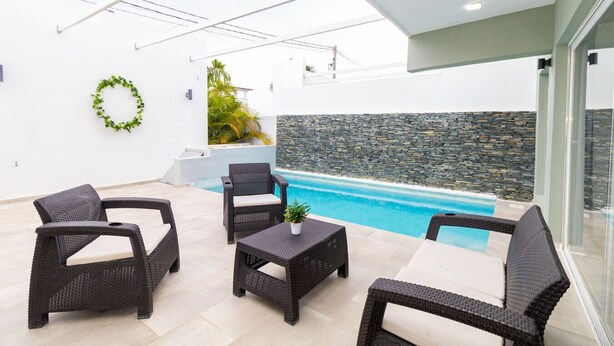 A modern outdoor patio with wicker furniture, a pool, and a stone wall. - Tropical 3BR Retreat w/ Private Pool in Bayahibe (San Rafael del Yuma)