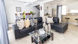 A modern living room with a black leather sofa, a glass coffee table, and a kitchen area in the background. - Tropical 3BR Retreat w/ Private Pool in Bayahibe (San Rafael del Yuma)