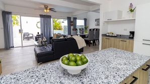 A modern living room with a black sofa, a dining area, and a kitchen with a granite countertop. - Tropical 3BR Retreat w/ Private Pool in Bayahibe (San Rafael del Yuma)
