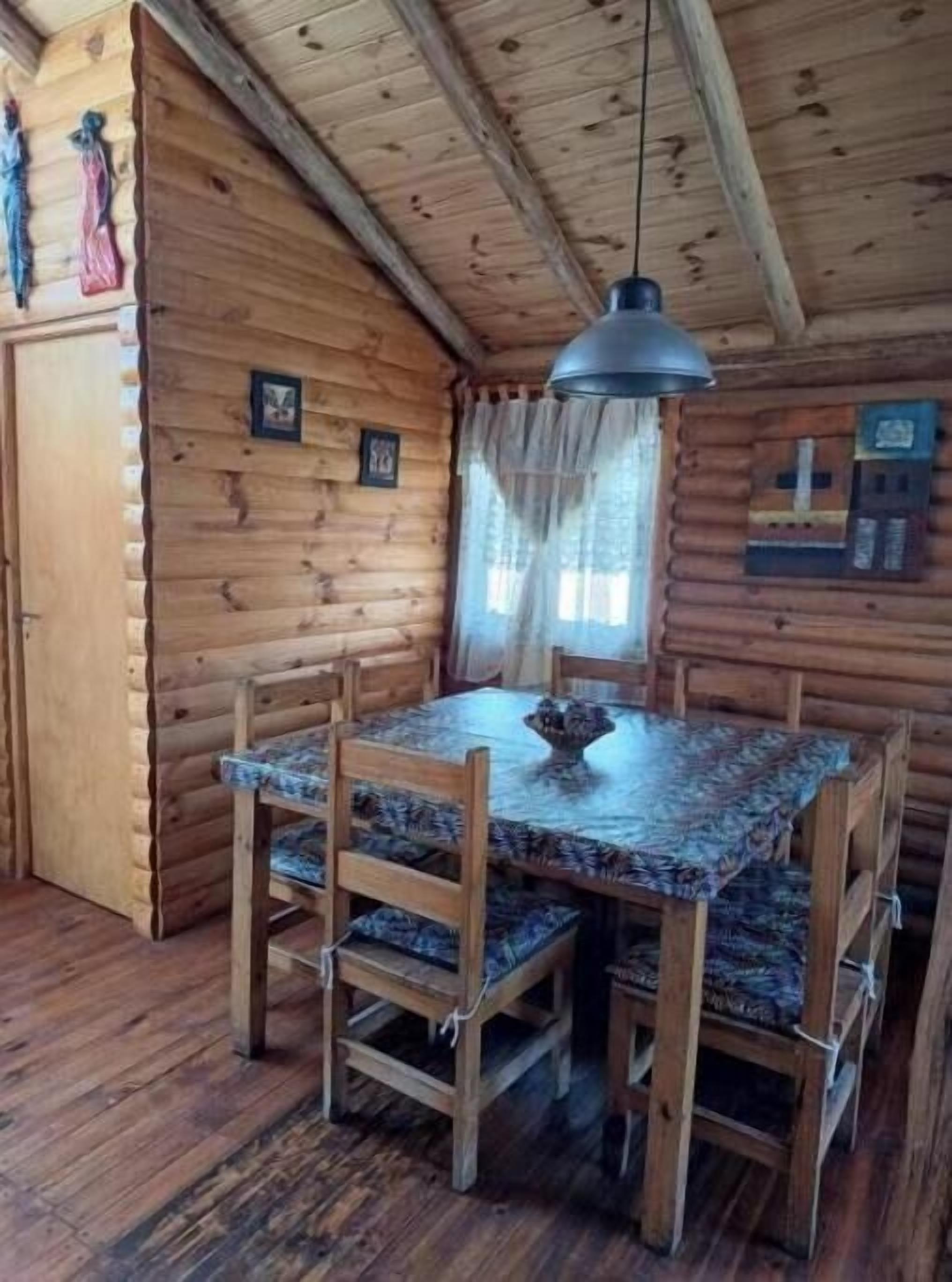 A wooden cabin interior with a dining table, chairs, a hanging light fixture, and a small statue on the wall.