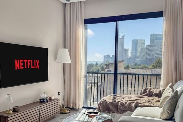 A modern living room with a large TV displaying "NETFLIX", a sofa, a coffee table, and a view of a cityscape through large windows. - Chic, cozy studio just 2 minutes from Anfa Park and CFC. (Casablanca)