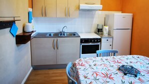 Cocina compacta con lavaplatos, estufa y refrigerador. Hay una silla azul y una mesa con mantel floreado.