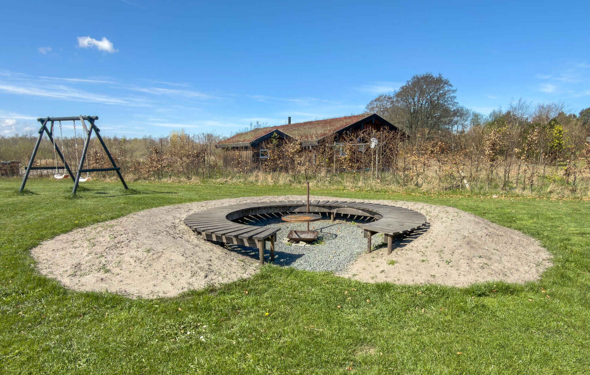 Ein runder Sitzbereich im Freien mit Feuerstelle, umgeben von einer Wiese und einer hölzernen Schaukelanlage.