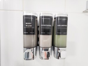 Drei Spender mit den Aufschriften 'BODY WASH', 'CONDITIONER' und 'SHAMPOO' in einem Badezimmer.