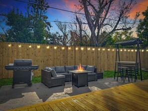 Ein Hinterhof mit einer Holzterrasse, einem Grill, einer Feuerstelle und Gartenmöbeln.