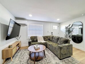 A living room with a sectional sofa, a coffee table, a TV mounted on the wall, and a large mirror. - Sunny Nokomis Getaway•Private Pool•Backyard Oasis (Nokomis)