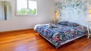 Une chambre avec un plancher en bois, un lit recouvert d’une literie à motifs floraux, une fenêtre laissant entrer la lumière du jour et un climatiseur fixé au mur.