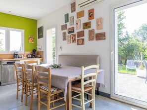 A room with a dining table, chairs, a kitchen area, and a large window with a view of greenery. - Maison Au Petit Bonheur (Argenton-sur-Creuse)