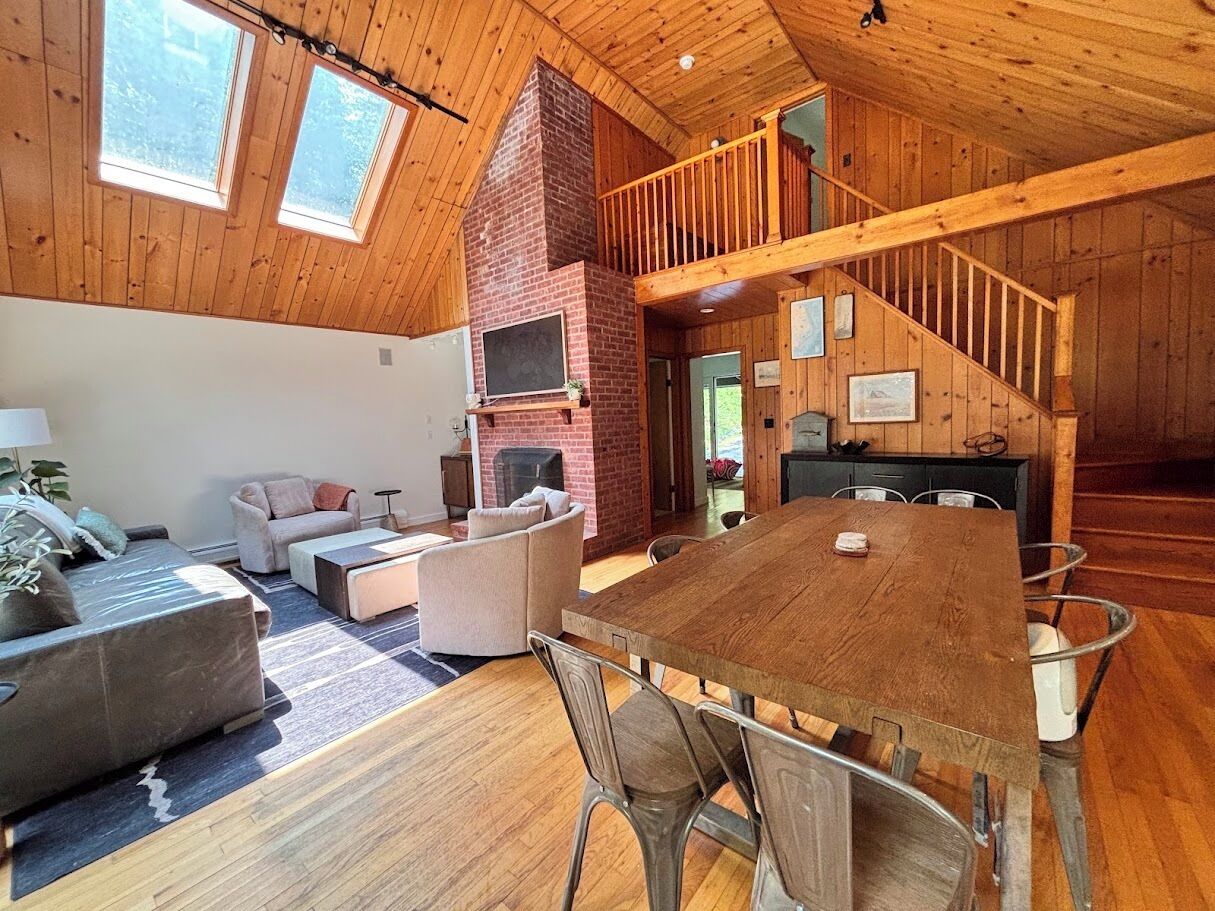 Un vaste espace de vie comprenant une grande table à manger en bois, des sièges confortables, une cheminée et un escalier menant à un niveau supérieur.