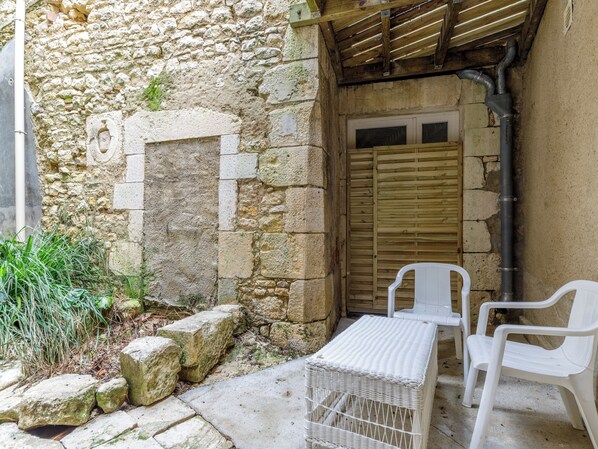 Un patio con paredes de piedra, una puerta de madera, una mesa de mimbre blanco y dos sillas de plástico blanco.