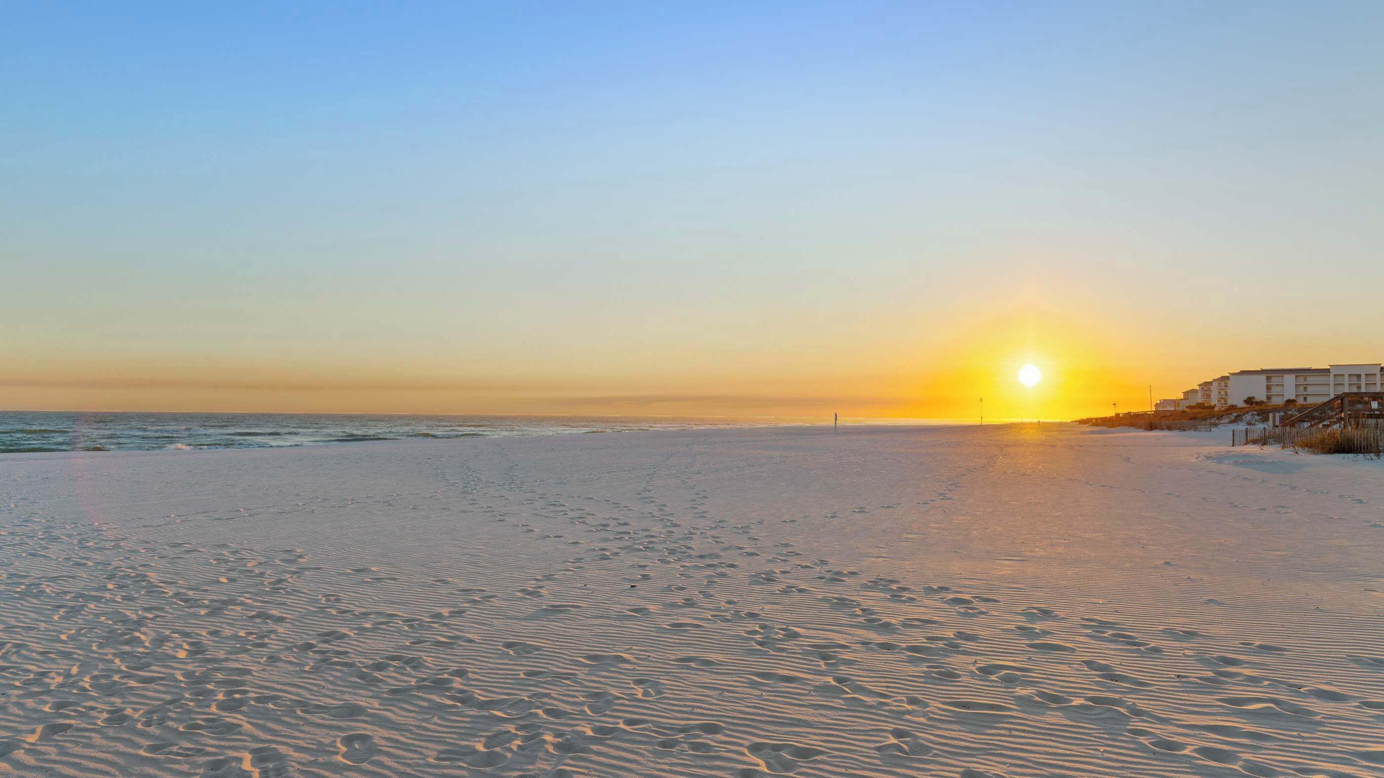 A beach with footprints leading to the water, a sunset in the background, and buildings on the right.