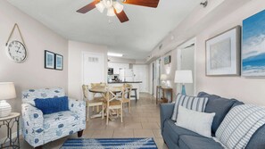 A living room with a blue patterned sofa, a grey sofa, a wooden dining table with chairs, a kitchen in the background, and a ceiling fan. - Condo With Gulf View Direct Beach Access, Indoor/outdoor Pool (Orange Beach)