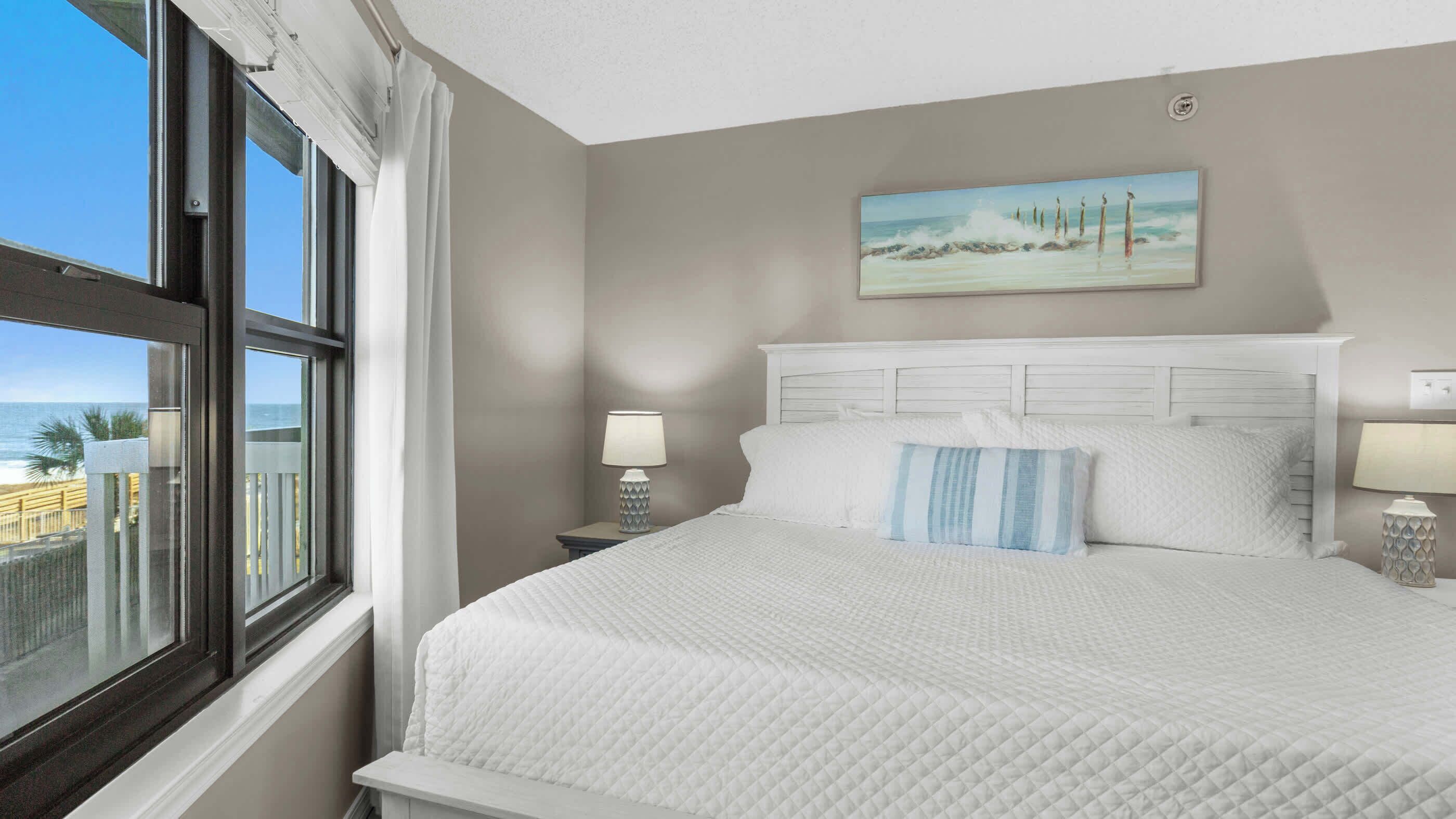 A hotel room with a large bed, a painting of a beach scene, and a view of the ocean through the window.