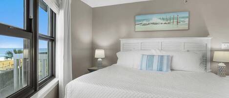 A hotel room with a large bed, a painting of a beach scene, and a view of the ocean through the window.