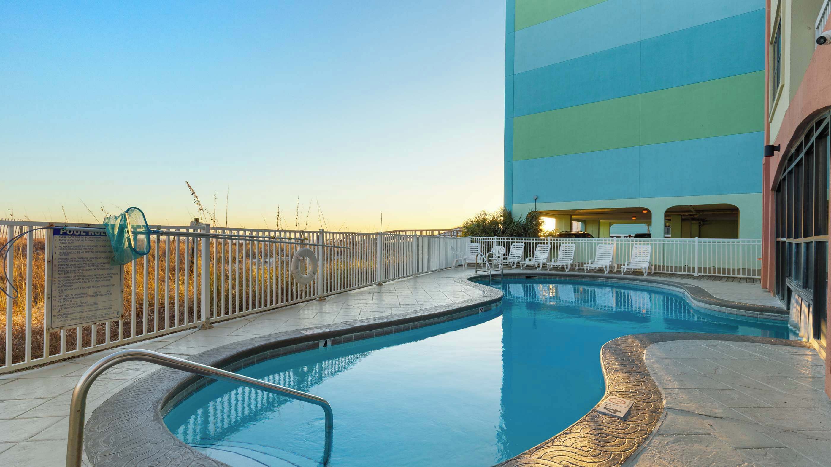 A swimming pool with a white fence, lounge chairs, and a building with a blue and green facade.