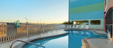 A swimming pool with a white fence, lounge chairs, and a building with a blue and green facade.