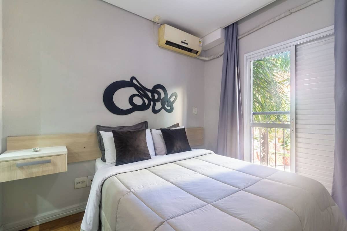 Une chambre à coucher comprenant un lit, une table de chevet, un appareil de climatisation et une fenêtre avec des rideaux.
