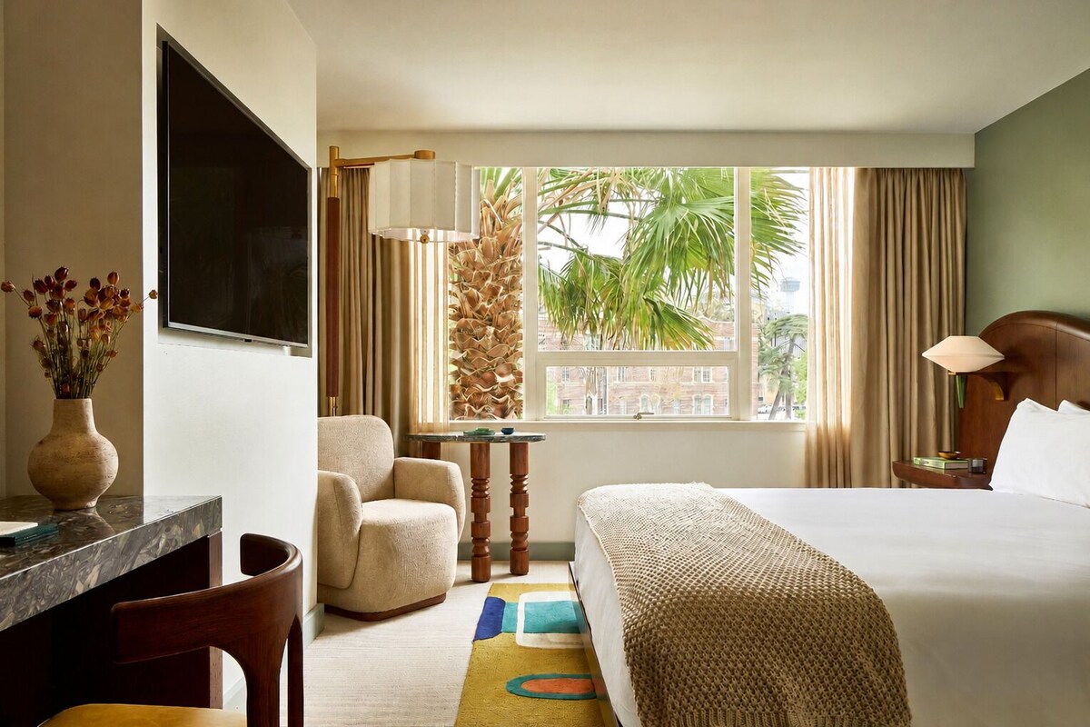 A hotel room with a bed, a desk, a chair, a TV, a window with curtains, and a view of palm trees.