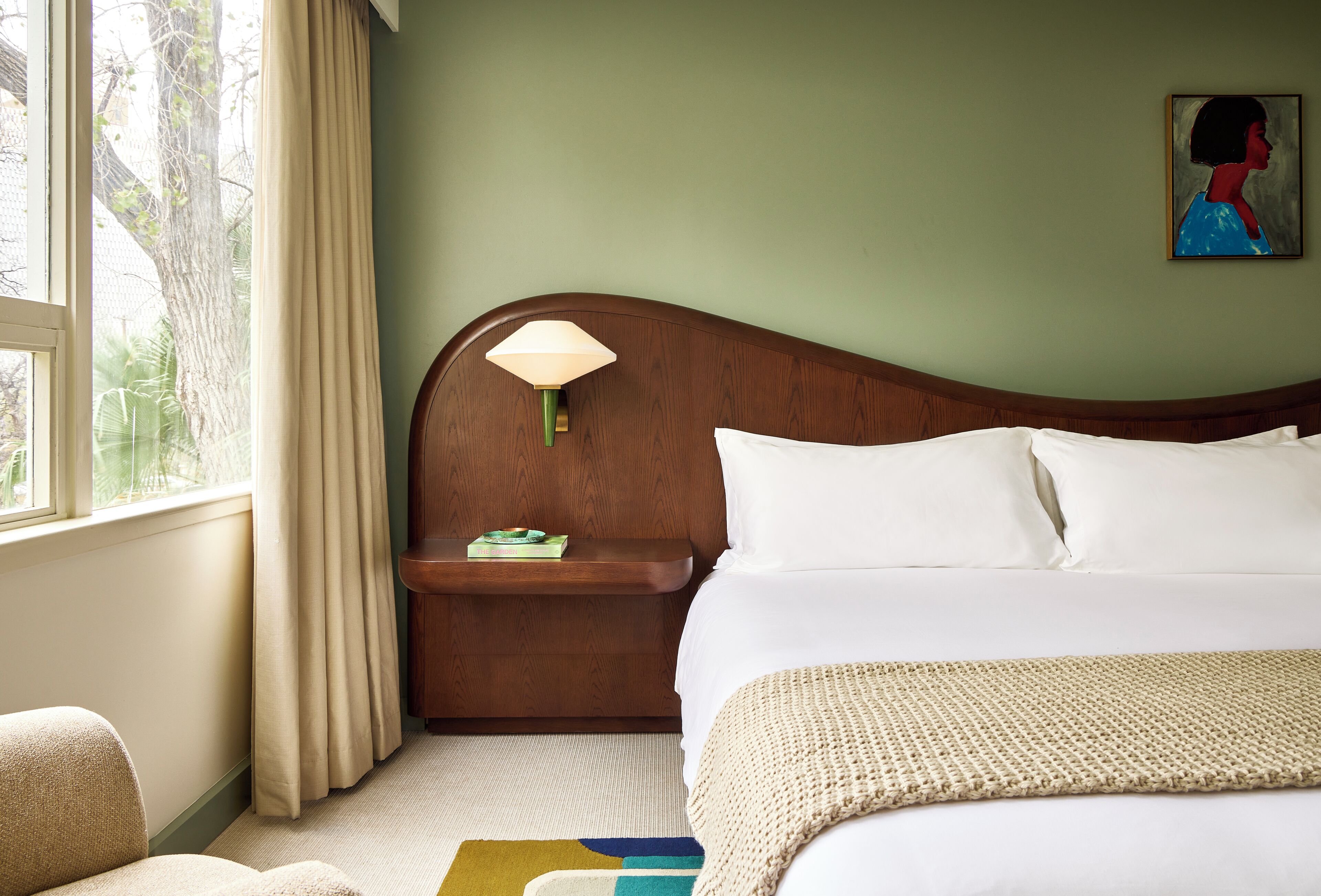 Una camera d'albergo con un letto, un comodino, una lampada, un quadro e vista su alberi dalla finestra.