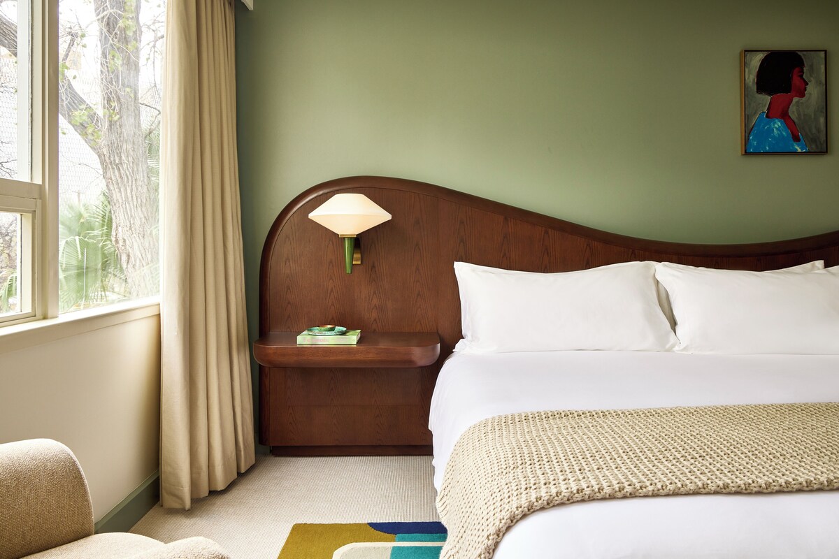 A hotel room with a bed, a nightstand, a lamp, a painting, and a view of trees through the window.