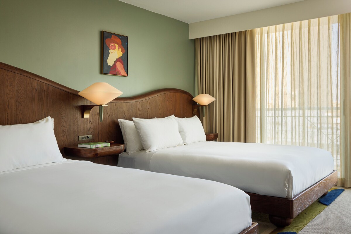 A hotel room with two beds, a wooden headboard, a painting on the wall, and a window with curtains.