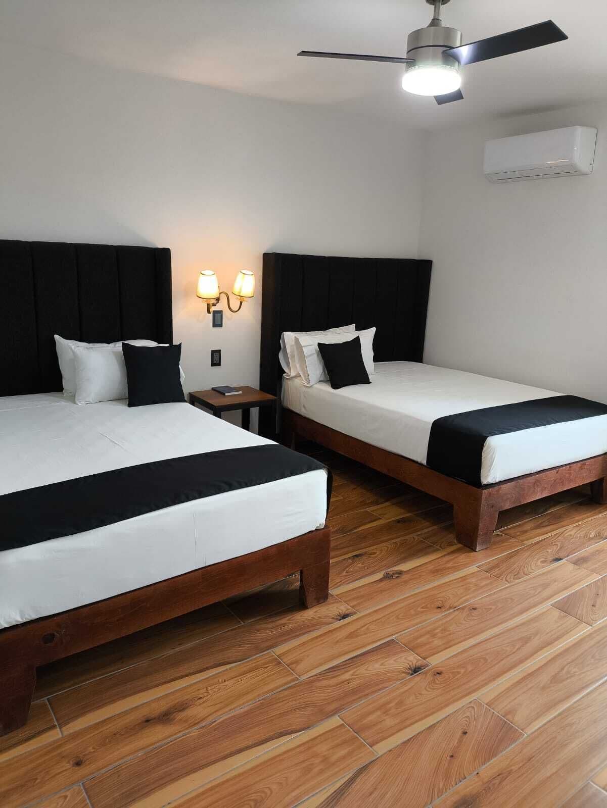 Habitación de hotel con dos camas, ventilador de techo, aire acondicionado de pared y mesita de noche con lámpara.