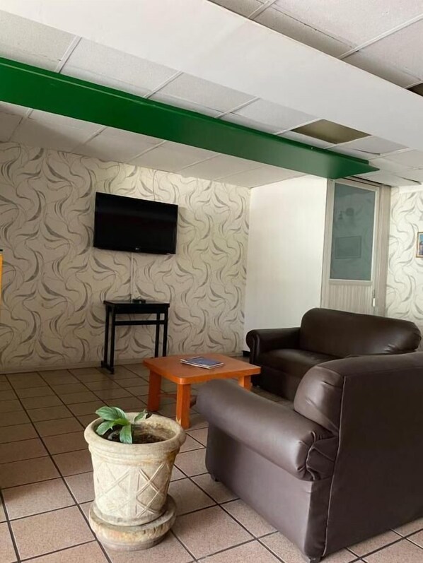 A living room with a brown leather sofa, a small wooden table, a potted plant, a television mounted on the wall, and patterned wallpaper. - Hotel Sicilia (Aguascalientes)