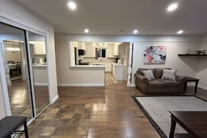 A spacious living area with a wooden floor, a sofa, and a coffee table. There is a kitchen in the background with white cabinets and a dining area visible through a glass door.
