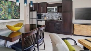Eine moderne Küche mit dunklen Holzschränken, einem Edelstahlkühlschrank, einer Mikrowelle und einem großen Flachbildfernseher. Es gibt einen Essbereich mit einem Holztisch und Stühlen sowie ein farbenfrohes abstraktes Gemälde an der Wand.