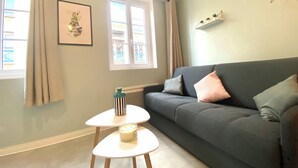 A living room with a dark grey sofa, two small round tables, a framed picture on the wall, and a window with curtains. - Le 202 : studio rénové au coeur de Rouen (Rouen)