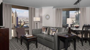 Habitación de hotel moderna con un sofá gris, una mesita y un área de comedor con sillas. Amplias ventanas ofrecen vistas de la ciudad.